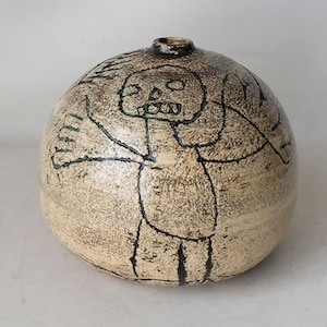 May include: A white ceramic vase with a round shape and a black and green drawing of a stick figure with a skull on its head.