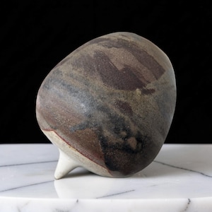 Op de afbeelding: Een handgemaakte keramische sculptuur met een unieke, organische vorm. Het stuk heeft een mix van aardetinten, waaronder bruin, grijs en hints van rood. Het heeft een kleine, witte, puntige basis en wordt gepresenteerd op een marmeren oppervlak.