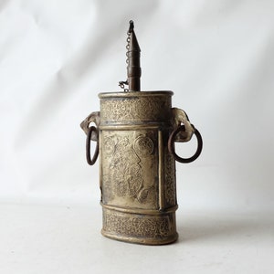 Op de afbeelding: Een antieke zilverkleurige metalen container met ingewikkelde gegraveerde ontwerpen. Het heeft twee metalen ringen aan de zijkanten en een tuit aan de bovenkant met een ketting.