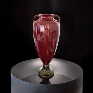 May include: A tall, burgundy glass vase with two handles near the top. The vase has a flared shape and sits on a dark green base. The vase is displayed on a black, cylindrical pedestal against a black background.