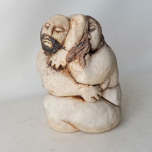 Op de afbeelding: Een keramische sculptuur van twee figuren die elkaar omhelzen. De figuren zijn naakt en hebben realistische kenmerken. De sculptuur is geschilderd in tinten bruin en wit.