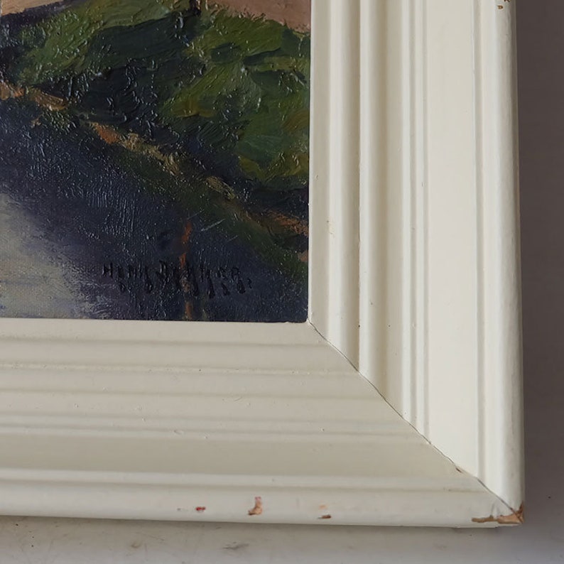 May include: A close-up of a white wooden frame with a painting inside. The painting is of a landscape with green trees and a light blue sky. The artist's signature is visible in the bottom right corner of the painting.