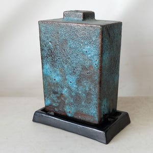 Puede incluir: Un jarrón de cerámica azul turquesa con una superficie texturizada y una base negra. El jarrón tiene una forma rectangular y una parte superior cuadrada.