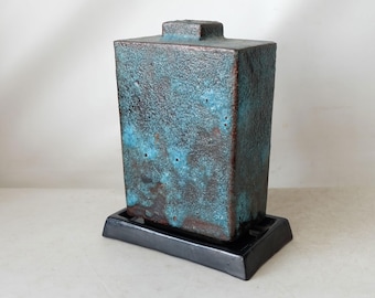 Puede incluir: Un jarrón de cerámica azul turquesa con una superficie texturizada y una base negra. El jarrón tiene una forma rectangular y una parte superior cuadrada.