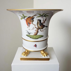 May include: White ceramic vase with a flared top and gold trim. The vase features a detailed illustration of two birds perched on a tree branch. The base has gold-colored claw feet and a gold trim. A small butterfly is also depicted.