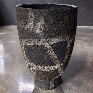 Puede incluir: Un jarrón alto y cónico con una superficie oscura, de color negro carbón, porosa y texturizada. El exterior presenta un patrón abstracto contrastante, rugoso y de color blanco roto, con gruesas líneas curvas.