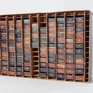 Könnte beinhalten: Eine Holzkiste mit einem Gitter aus kleinen Fächern, die jeweils einen kleinen, rechteckigen, braunen Gegenstand enthalten. Die Gegenstände sind mit weißer Schrift nummeriert, einige haben rote Schrift. Die Nummern reichen von 271 bis 386.