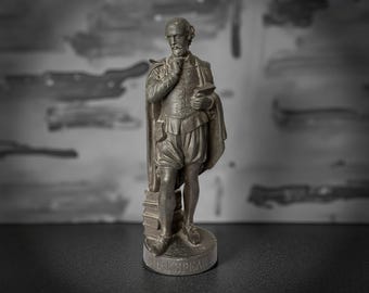 May include: A bronze-colored statue of William Shakespeare, holding a book and with his hand on his chin. The statue is on a round base with the name "SHAKESPEARE" inscribed. The figure is dressed in period clothing.