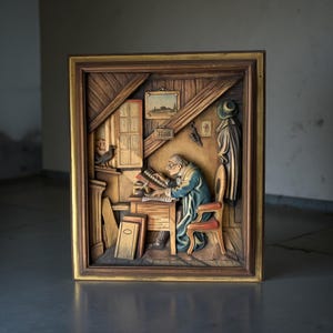 Puede incluir: Un relieve de madera tridimensional enmarcado que representa a un hombre sentado en un escritorio en una habitación rústica. El hombre lleva un abrigo azul y trabaja en un libro. El relieve está en un marco de madera con un borde dorado.