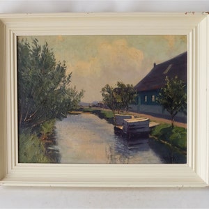 May include: A framed oil painting depicting a rural scene with a canal, a house with a thatched roof, and trees. The sky is a pale blue with white clouds. The painting is in a white frame.