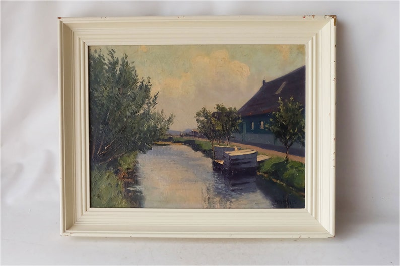 May include: A framed oil painting depicting a rural scene with a canal, a house with a thatched roof, and trees. The sky is a pale blue with white clouds. The painting is in a white frame.