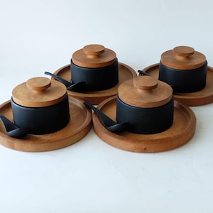 Op de afbeelding: Vier zwarte keramische kommen met houten deksels en bijpassende houten voetjes. Elke kom heeft een klein zwart lepeltje.