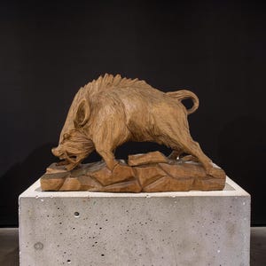 May include: A detailed wooden sculpture of a boar in a charging pose. The boar is carved with intricate detail, showcasing its bristly fur and open mouth. The sculpture is mounted on a carved wooden base, and is displayed on a concrete plinth.