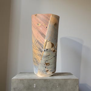 Könnte beinhalten: Eine hohe, abstrakte Keramikvase mit einem einzigartigen, kantigen Design. Die Vase weist eine Mischung aus Farben auf, darunter Rosa, Beige und Blau, mit schwarzen und braunen abstrakten Mustern. Sie steht auf einem grauen Betonsockel und zeigt ihre künstlerische Form.