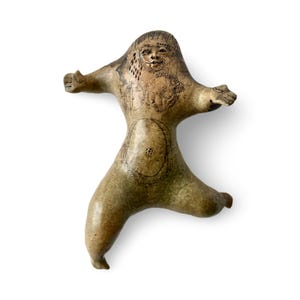 May include: A ceramic figurine in a standing pose with arms outstretched. The figure has a smiling face and intricate etched details on the body, including patterns on the chest and abdomen. The color is a muted, earthy tone.