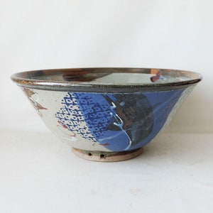 Peut inclure: Un grand bol en céramique avec un glaçage blanc et un motif abstrait bleu, brun et blanc. Le bol a un bord légèrement évasé et un petit pied.