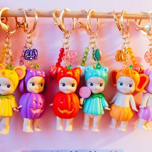 May include: Six keyrings with small doll figures in Halloween costumes, including a mouse, pumpkin, and frog. Each figure holds a lollipop and hangs from a gold-coloured chain and ring. The keyrings are suspended from a wooden rod.