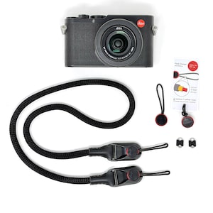 May include: A black Leica digital camera with a lens, a black braided camera strap with two quick-release connectors, two additional circular anchor links, two small metal attachment rings, and a Peak Design instruction card with red text.