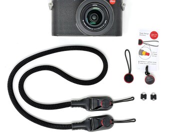 Op de afbeelding: Een zwarte Leica digitale camera met lens, een zwarte gevlochten camerariem met twee snelkoppelingen, twee extra ronde ankerlinks, twee kleine metalen bevestigingsringen en een Peak Design instructiekaart met rode tekst.