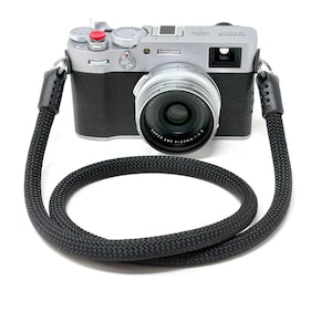 May include: A silver Fujifilm X100V digital camera with a black textured grip and a red shutter button. The camera features a Super EBC f=23mm 1:2 II lens and is attached to a thick, black braided rope camera strap with black leather connectors.