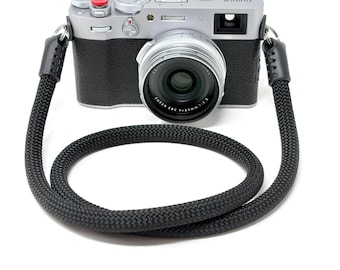 Könnte beinhalten: Eine silberne Fujifilm X100V Digitalkamera mit schwarzem, texturiertem Griff und rotem Auslöser. Die Kamera verfügt über ein Super EBC f=23mm 1:2 II Objektiv und ist an einem dicken, schwarzen geflochtenen Seil-Kameragurt mit Lederenden befestigt.