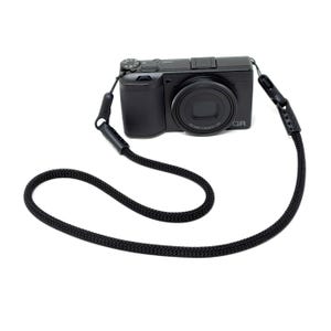 May include: A black digital camera with a fixed lens and the text GR printed on the front, attached to a thick, braided black rope neck strap. The strap features black leather-like connectors at both ends that attach to the camera body.