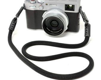 Könnte beinhalten: Eine silberne Fujifilm X100V Digitalkamera mit schwarzem, strukturiertem Griff und einem roten Auslöser. Die Kamera ist an einem dicken, schwarzen geflochtenen Seil-Trageriemen befestigt, der an beiden Enden des Kameragehäuses schwarze Lederverbinder aufweist.