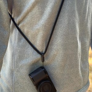 Pode incluir: Uma câmara digital preta com lente e as letras GR impressas na frente, presa a uma correia de pescoço de cordão entrançado preto. A câmara está pendurada na correia, posicionada contra o tecido de uma camisola desportiva cinzenta.