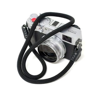 May include: A silver Fujifilm X100V digital camera with a black woven camera strap. The camera has a silver lens, a red button, and black leather accents. The strap is looped around the camera.