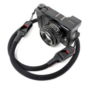 Puede incluir: Cámara Lumix negra con una lente negra y una correa de cámara negra tejida. La correa tiene costuras rojas y un botón rojo. La cámara está sobre un fondo blanco.