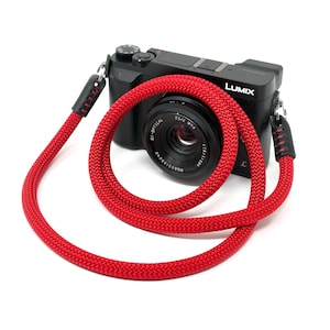 May include: A black Lumix digital camera with a bright red rope camera strap. The strap is looped around the camera and has black leather accents at the ends. The camera lens is visible.