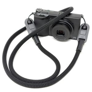 May include: Black camera with a black woven neck strap. The camera has a lens and several buttons on top. The strap has grey adjustable connectors. The camera is labelled "GR" on the front.