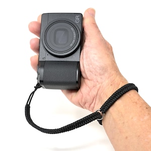Puede incluir: Una cámara digital negra con una correa de muñeca se sostiene en una mano. La cámara tiene una lente grande y las letras "GR" son visibles. La correa de muñeca es negra y trenzada, con un anillo metálico para sujetarla.