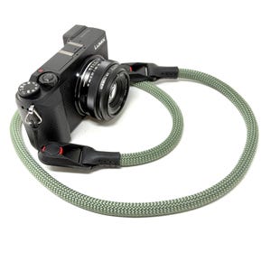 Puede incluir: Cámara digital negra con lente y correa de cámara de cuerda verde y gris. La correa de la cámara está sujeta a la cámara en ambos lados con clips de plástico negro. La cámara está sobre un fondo blanco.