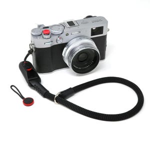 Puede incluir: Una cámara plateada con una lente negra y una correa de cámara negra con detalles rojos. La cámara tiene un botón rojo en la parte superior.