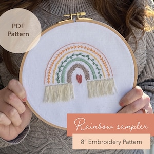 May include: An 8-inch embroidery pattern featuring a colorful rainbow design. The rainbow includes shades of yellow, green, and brown, with a heart at the center. The design is framed in a wooden hoop, with the text "Rainbow sampler" and "PDF Pattern".
