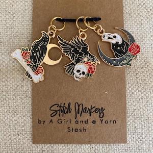 May include: Three stitch markers with black raven, skull, and bone designs, accented with red roses and gold-colored metal. The markers are attached to a brown card with the text "Stitch Markers by A Girl and a Yarn Stash."