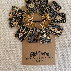 May include: A set of 10 gold stitch markers shaped like tarot cards with black and gold designs. The cards include The Lovers, The Strength, The Magician, The Moon, The Sun, Death, The Wheel of Fortune, and more. The stitch markers are attached to a ring and are labeled "Stitch Markers by A Girl and a Yarn Stash."