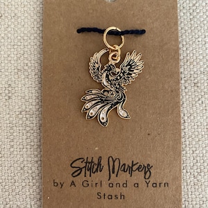 May include: A gold-colored stitch marker with a black and white phoenix design. The stitch marker is attached to a brown card with the text "Stitch Markers by A Girl and a Yarn Stash."