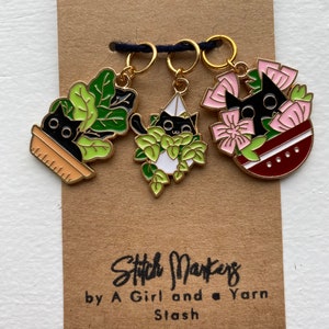 May include: Three gold stitch markers with black cats in different settings. The first marker features a black cat in a pot of green plants. The second marker features a black cat in a hanging planter with green plants. The third marker features a black cat in a pink flower pot with red and white flowers. The markers are labeled "Stitch Markers by A Girl and Yarn Stash."