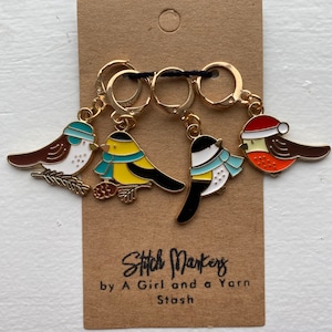 May include: Set of four gold-colored stitch markers featuring bird designs. Each bird has a unique color scheme, including brown, yellow, black, and orange, with accents of blue and white. The markers are attached to a brown card with the text "Stitch Markers by A Girl and a Yarn Stash."
