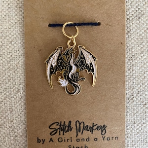 May include: Gold stitch marker featuring a black and white dragon with stars on its wings. The text "Stitch Markers by A Girl and a Yarn Stash" is printed on the brown card.