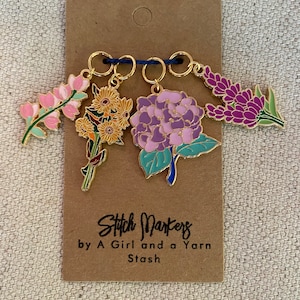 Puede incluir: Cuatro marcadores de punto florales con anillos dorados e hilo azul. Las flores incluyen flores rosas, amarillas, moradas y lavanda. La tarjeta dice "Stitch Markers by A Girl and a Yarn Stash."
