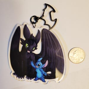 May include: A clear acrylic keyring featuring a printed illustration of a black dragon with green eyes and a small blue alien creature. The keyring includes a black metal bat-shaped clasp and is shown next to a standard US quarter for scale.