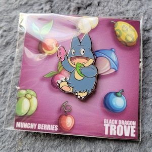 Puede incluir: Un pin con un personaje de dibujos animados azul que sostiene un objeto verde y una fruta rosa. El pin está sobre un fondo púrpura con varias frutas coloridas y el texto "Munchy Berries" y "Black Dragon Trove".