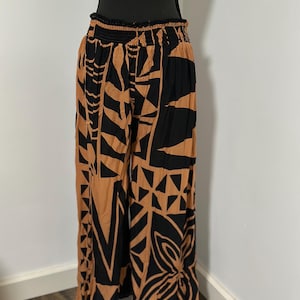May include: Wide-leg pants with a black and tan geometric pattern. The pants have a gathered waistband and a loose, flowing fit. The design features abstract shapes and floral motifs, creating a bold and stylish look. The pants are displayed on a mannequin.