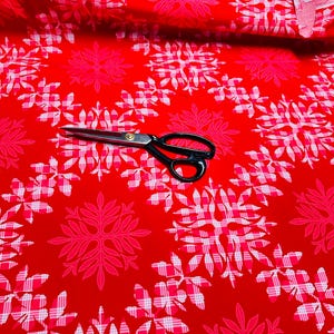 May include: Red fabric with a repeating pattern of white and pink floral and plaid designs. A pair of black-handled scissors rests on the fabric, suggesting it's for sewing or crafting projects.