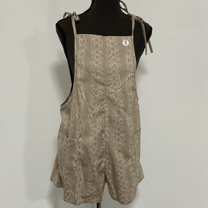 Peut inclure: Une salopette en jean beige avec un motif floral blanc. La salopette a des bretelles r&eacute;glables et deux poches. L'&eacute;tiquette de taille indique "S".