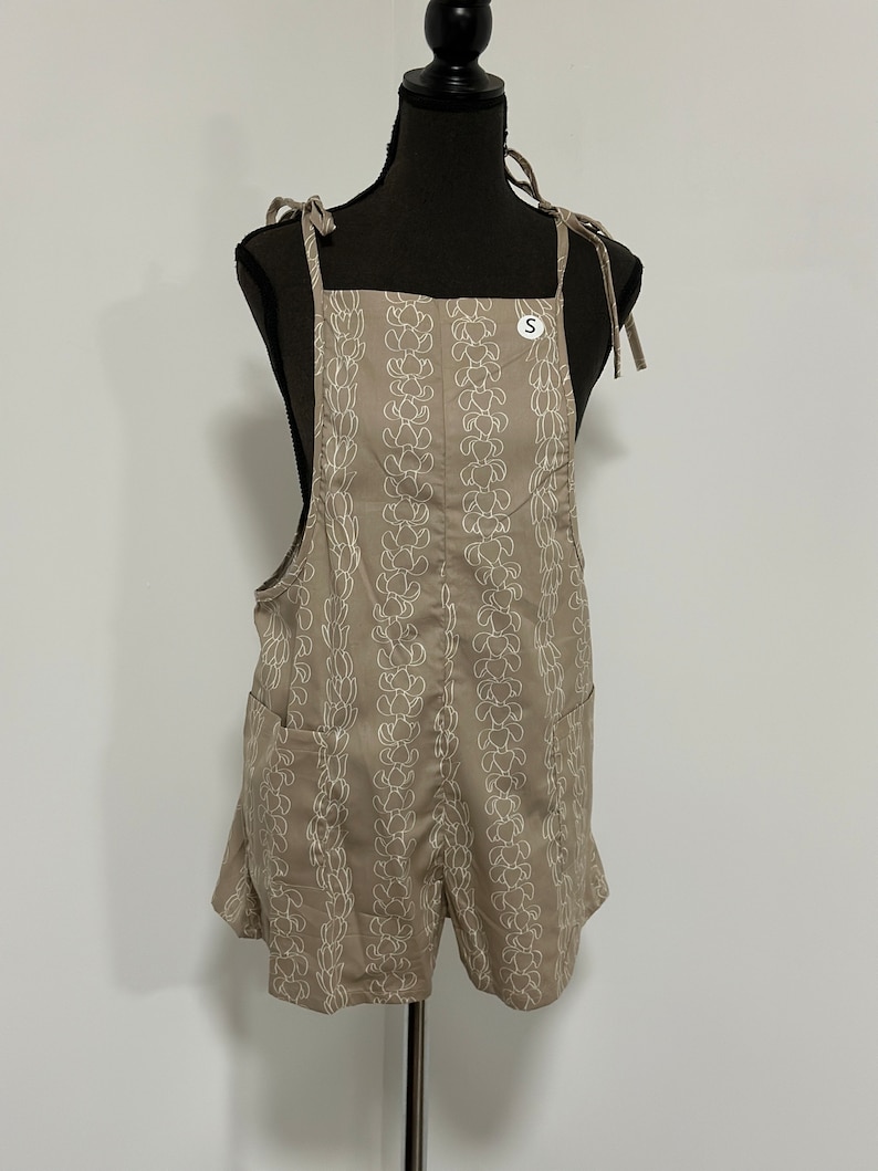 Peut inclure: Une salopette en jean beige avec un motif floral blanc. La salopette a des bretelles r&eacute;glables et deux poches. L'&eacute;tiquette de taille indique "S".