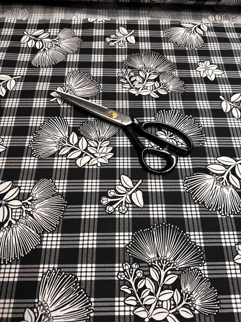 May include: Black and white floral fabric with a plaid pattern. The fabric features white botanical illustrations on a black background. A pair of black handled scissors with a gold center are on top of the fabric.